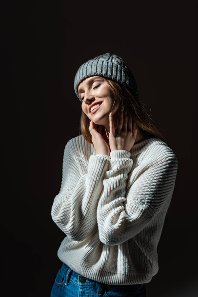 beautiful dreamy tender girl with closed eyes in sweater and hat isolated on black - Foto, imagen