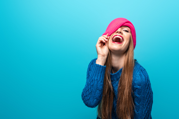 attractive funny woman laughing in knitted sweater and pink hat, isolated on blue - Φωτογραφία, εικόνα