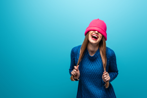 funny laughing woman in knitted sweater with pink hat on eyes, isolated on blue - Photo, Image