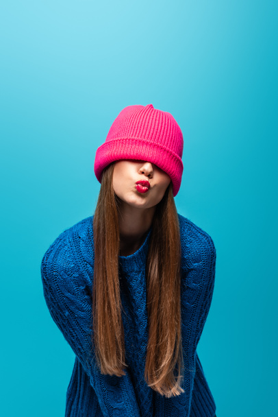 attractive woman in knitted sweater with pink hat on eyes making air kiss, isolated on blue - Photo, image