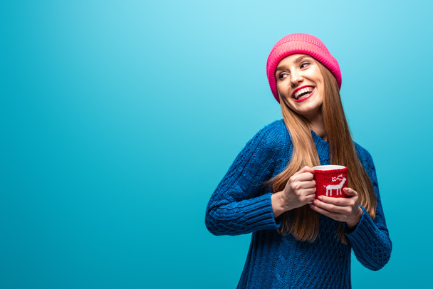 attractive happy woman in knitted sweater and pink hat holding cup of coffee, isolated on blue - Foto, Imagem