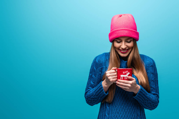 smiling woman in knitted sweater and pink hat holding cup of coffee, isolated on blue - Photo, Image
