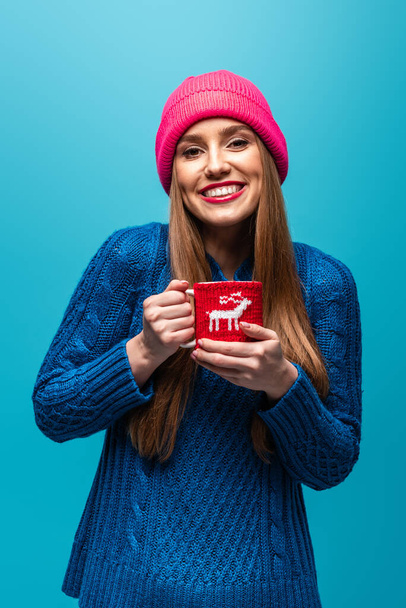 beautiful woman in knitted sweater and pink hat holding cup of coffee, isolated on blue - Photo, Image