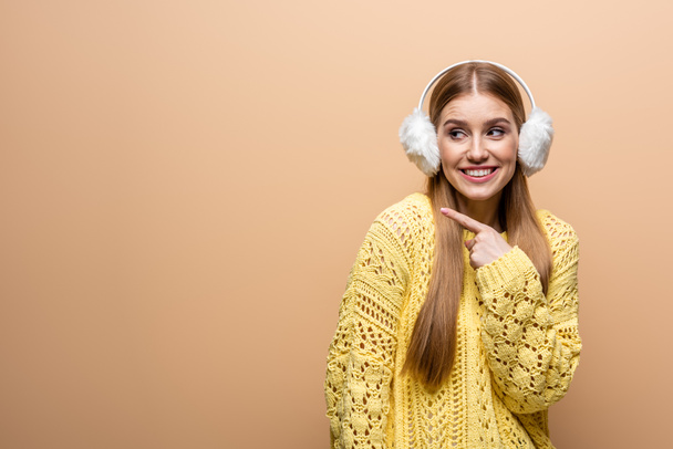 beautiful smiling woman pointing in yellow sweater and earmuffs, isolated on beige - Foto, immagini