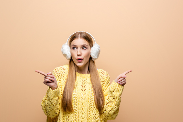 beautiful shocked girl in yellow sweater and earmuffs pointing at something, isolated on beige - Фото, зображення
