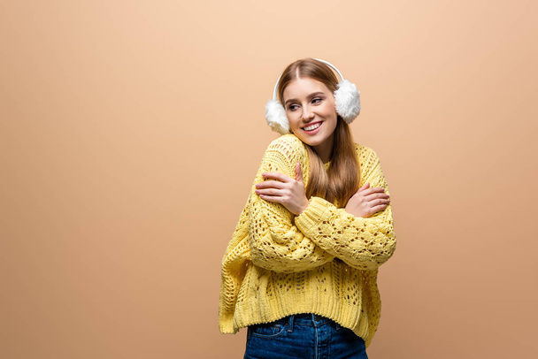 koude lachende vrouw in gele trui en oorwarmers, geïsoleerd op beige - Foto, afbeelding