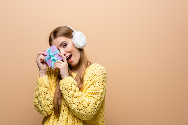 excited woman in yellow sweater and earmuffs holding christmas present, isolated on beige - Photo, Image