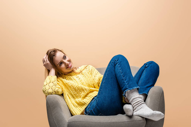 atraktivní usměvavá žena ve žlutém svetru sedící na křesle, izolovaná na béžové - Fotografie, Obrázek