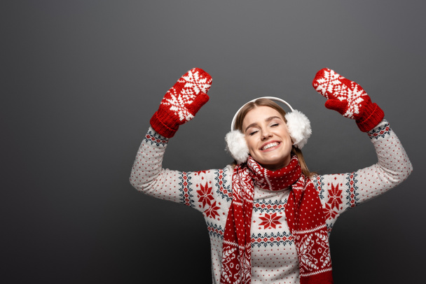 atrakcyjna wesoła dziewczyna w świątecznym swetrze, szaliku, rękawiczkach i nausznikach, odizolowana na szaro - Zdjęcie, obraz