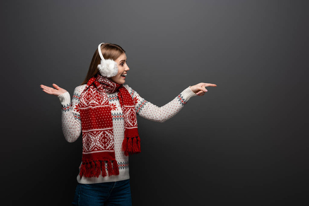 excited woman in christmas sweater, scarf and earmuffs pointing at something, isolated on grey - Фото, изображение