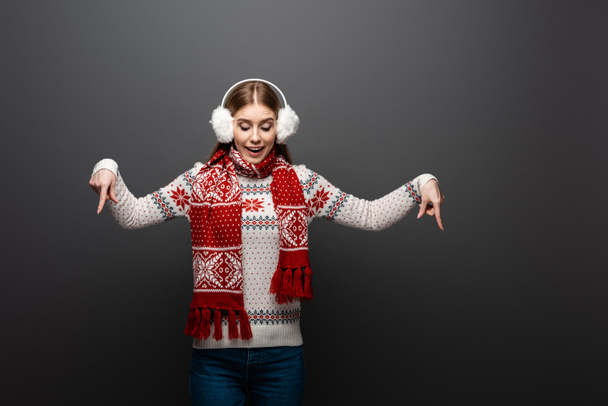 attractive girl in christmas sweater, scarf and earmuffs pointing down, isolated on grey - Photo, Image