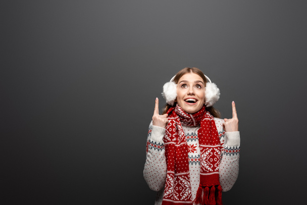 ελκυστική χαμογελαστή γυναίκα σε χριστουγεννιάτικο πουλόβερ, κασκόλ και ωτοασπίδες που δείχνουν προς τα πάνω, απομονωμένη σε γκρι - Φωτογραφία, εικόνα