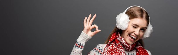 panoramatický záběr šťastné ženy ve vánočním svetru, šála a sluchátka mrkání a ukazování ok znamení, izolované na šedé - Fotografie, Obrázek
