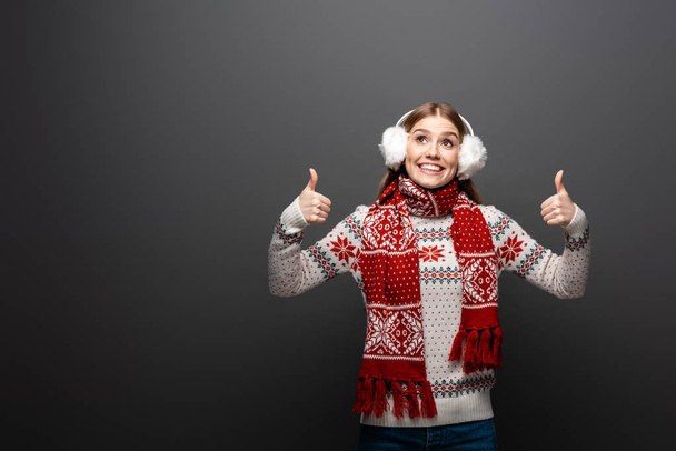 χαρούμενο κορίτσι με χριστουγεννιάτικο πουλόβερ, μαντήλι και ωτοασπίδες να κλείνει το μάτι και να δείχνει τους αντίχειρες επάνω, απομονωμένο σε γκρι - Φωτογραφία, εικόνα