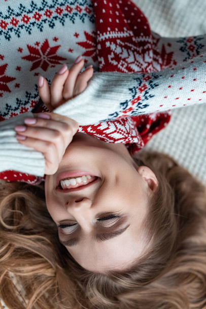 cheerful woman with closed eyes in warm sweater and scarf lying on blanket - Фото, зображення