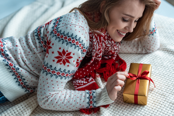 Ευτυχισμένη γυναίκα σε ζεστό πουλόβερ κρατώντας κουτί δώρου και ξαπλωμένη σε κουβέρτα - Φωτογραφία, εικόνα