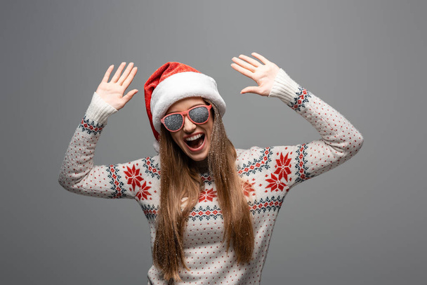 excited woman in christmas sweater, santa hat and sunglasses, isolated on grey - Photo, Image