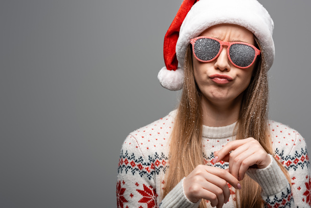 skeptical girl in christmas sweater, santa hat and sunglasses, isolated on grey - Zdjęcie, obraz