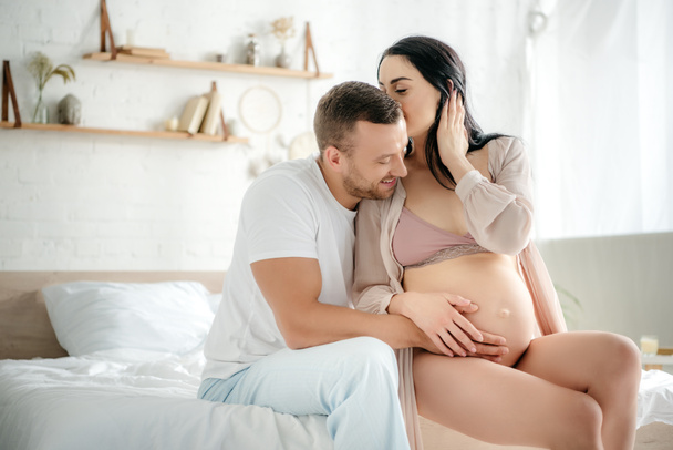 glücklicher Ehemann umarmt seine schwangere Frau, während sie ihn im Bett küsst - Foto, Bild