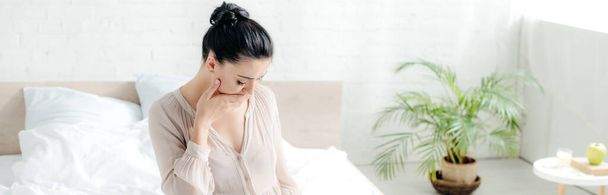 panoramic shot of beautiful pregnant girl in nightie having nausea while sitting on bed - Photo, image