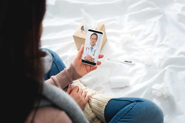 καλλιεργημένη άποψη της άρρωστης γυναίκας με χάπια και θερμόμετρο έχοντας σε απευθείας σύνδεση διαβούλευση με τον αρσενικό γιατρό στο smartphone - Φωτογραφία, εικόνα