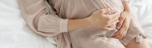 részleges kilátás terhes lány hálóingben megérinti has, miközben pihen az ágyon, panoráma lövés  - Fotó, kép