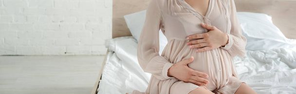 ベッドに座ってパノラマ撮影をしながらお腹に触れるドレスの妊婦のクロップドビュー  - 写真・画像