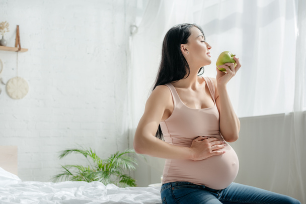 beautiful pregnant woman holding apple and touching belly while sitting on bed  - Foto, Imagen