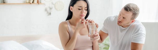 panorámica de la esposa con dolor tomar píldora con agua en el dormitorio con el marido cerca
 - Foto, Imagen