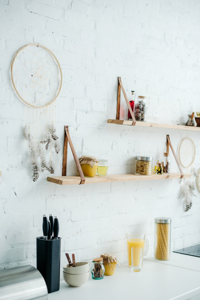 dream catchers and wooden shelves with jars on white brick wall in kitchen - Valokuva, kuva