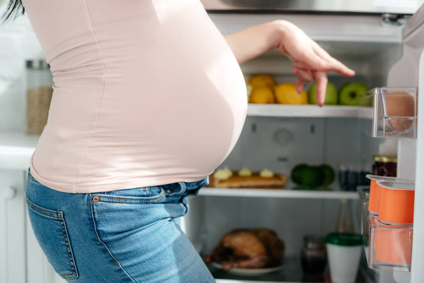 cropped view of pregnant woman pointing into opened fridge in kitchen - Photo, Image