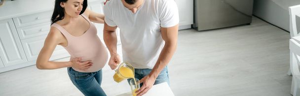 panoramiczne ujęcie pięknej ciężarnej żony w kuchni z mężem wylewającym sok pomarańczowy  - Zdjęcie, obraz
