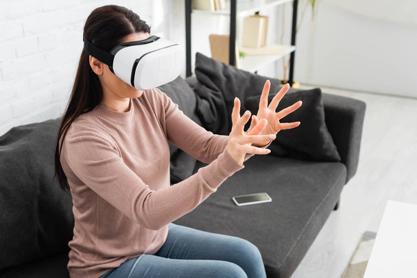 emotional woman using Virtual reality headset while sitting on sofa with smartphone - Foto, imagen