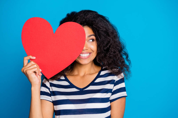 Close up photo of funky cheerful mulatto girl hold big red papercard heart close cover face look romantic boyfriend celebrate 14-february holiday wear striped t-shirt isolated blue color background - Foto, Bild