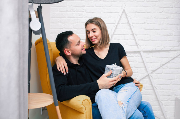 Portrait of couple young in a cozy room. A man sits on a yellow chair. A woman sits on the lap of a man and holds a gift - Foto, imagen