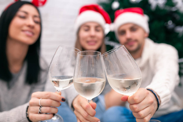 Happy friends celebrating New Year in home interior in Christmas hats sitting near a Christmas tree with glasses of wine. Wine glasses are the focus. Blurred figures of people behind glasses - 写真・画像