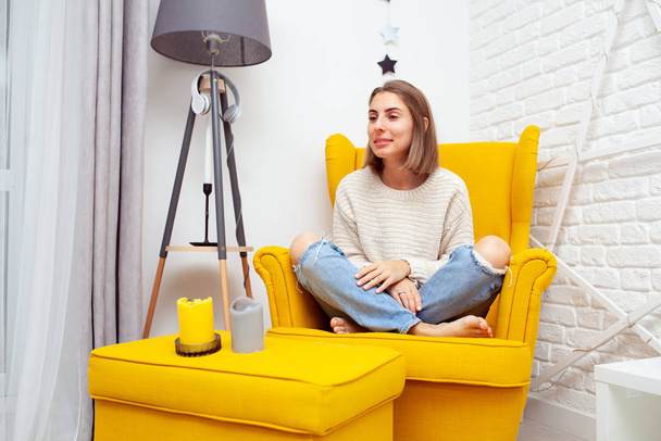 Enjoying leisure time at home. Beautiful young woman sitting in a soft yellow chair resting. Aromatic candles stand on the side - Valokuva, kuva