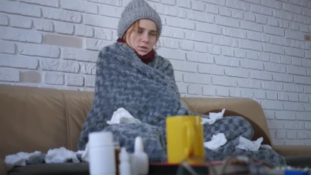 Covered with grey plaid young caucasian woman in hat and scarf freezing feeling cold at home, ill sick girl having fever flu influenza temperature symptoms wrapped in blanket shivering indoors. - Imágenes, Vídeo