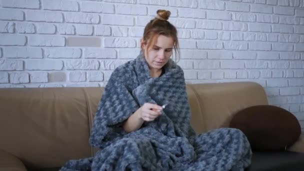 A fiatal kaukázusi nő rosszul érzi magát a kanapén ülve, majd lázmérővel ellenőrzi a testhőmérsékletet. Betegség személy betegség vírus portré.  - Felvétel, videó