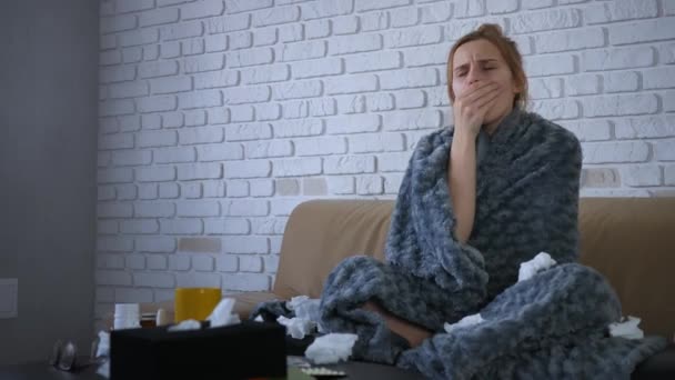 Beautiful young caucasian woman is sick, sitting in sofa. Her nose is stuffy, she sneezes and coughs. Blows nose and hides under the grey covers. - Footage, Video