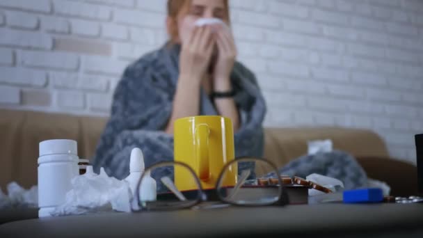 Young caucasian woman is sick, sitting in sofa, her nose is stuffy, she sneezes and coughs, blows nose and hides under the covers in defocus. In focus glasses medicine, pills, cup with tea.  - Footage, Video