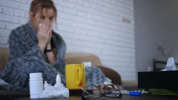 A fiatal kaukázusi nő beteg, a kanapén ül, az orra dohos, tüsszent és köhög, orrát fújja és elrejtőzik a takaró alatt. Fókuszban szemüveg orvoslás, tabletták, csésze tea.  - Felvétel, videó