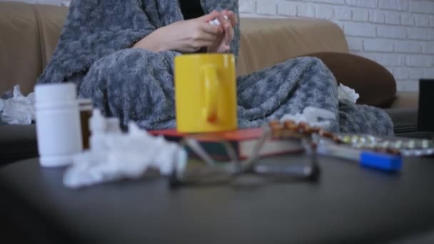 Krásná mladá běloška s kapajícím nosem používá nosní sprej doma, sedí na pohovce v obývacím pokoji pod přikrývkou - Záběry, video