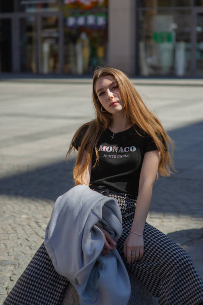 Een jong meisje met een zwart T-shirt en een grijze broek is op straat gefotografeerd. Mooie vrouw op een wandeling in de stad - Foto, afbeelding