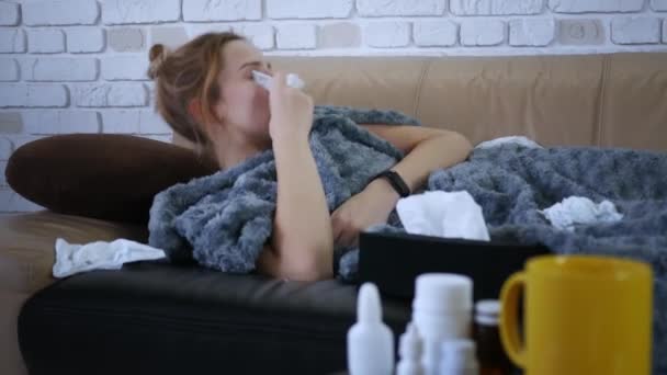 Hermosa mujer caucásica joven con secreción nasal utiliza un aerosol nasal en casa, acostado en el sofá en la sala de estar debajo de una manta
 - Metraje, vídeo