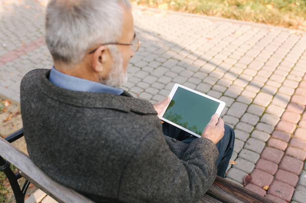 Se burlan de un hombre mayor usando tableta afuera. Vista posterior del anciano sentado en el banco y usando la tableta
 - Foto, Imagen