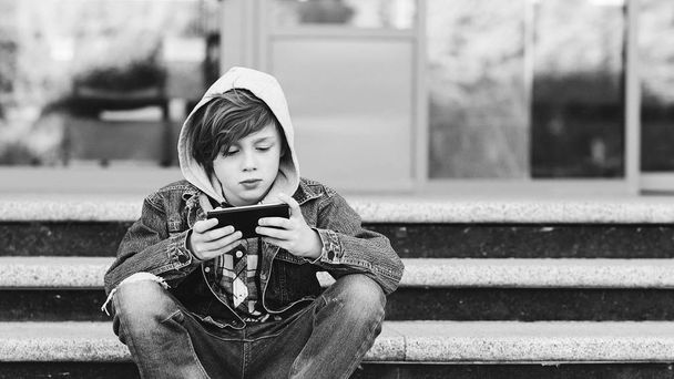 屋外の階段にスマートフォンを持つ10代。インターネット中毒の概念。少年は携帯電話でモバイルゲームをプレイ. - 写真・画像