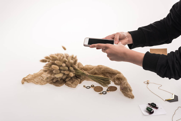 Ausgeschnittene Ansicht eines Fotografen, der Kompositionen mit Flora und Schmuck auf einem Smartphone auf Weiß fotografiert - Foto, Bild