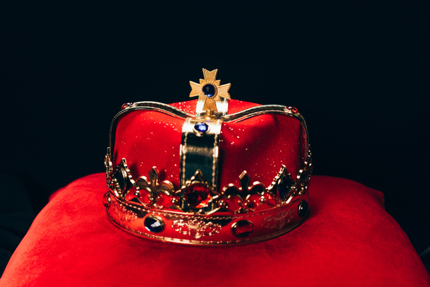 ancient golden crown with gemstones on red pillow, isolated on black - 写真・画像