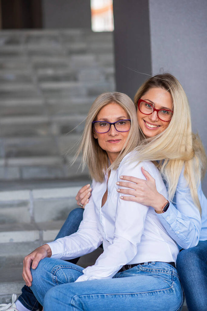 A beautiful blonde with glasses and her mature mother are sitting on the stairs outdoor. A well-groomed elderly woman hugs her adult daughter tightly. Love and family values. Mothers Day. - Photo, Image